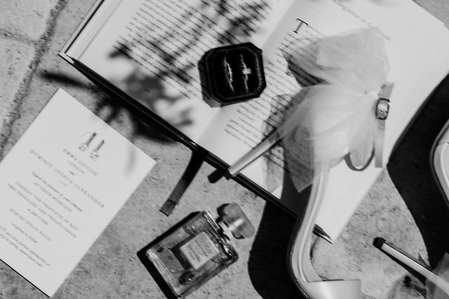 A black-and-white still life of a wedding menu, a ring box, perfume, and an open book on a table.