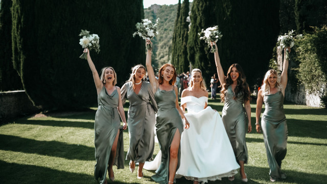 A joyful bride and her bridesmaids cheer and raise their bouquets on a sunlit lawn.