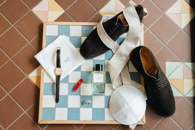 Flat-lay met zwarte nette schoenen, een horloge, een pochet en een stropdas op een betegeld dienblad.