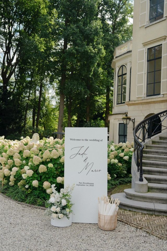 A white welcome sign with the couple's names set among white hydrangeas beside a mansion's curved staircase.