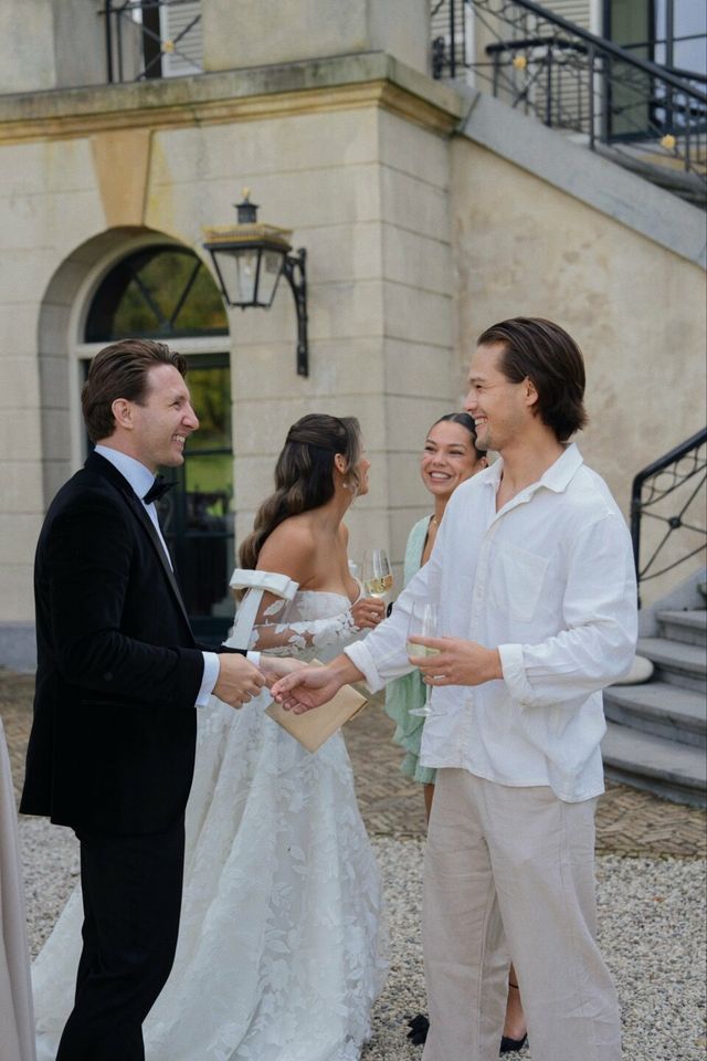 The groom in a tuxedo greets and shakes hands with a guest while the bride smiles beside the venue steps.