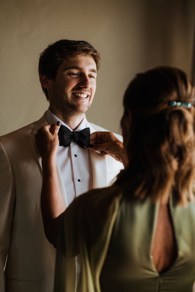 Groom in an ivory jacket having his black bow tie adjusted while getting ready