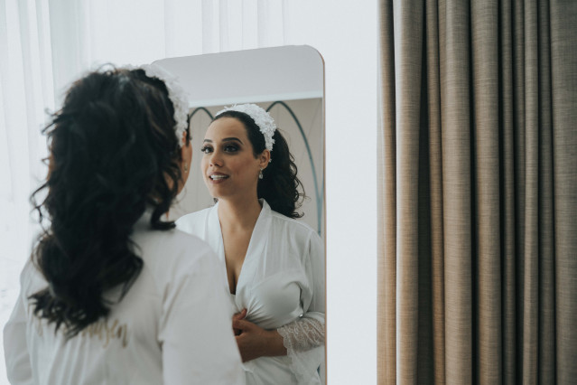 Bruid in witte badjas bekijkt haar opgemaakte gezicht in een staande spiegel tijdens de voorbereiding op een hotelkamer.