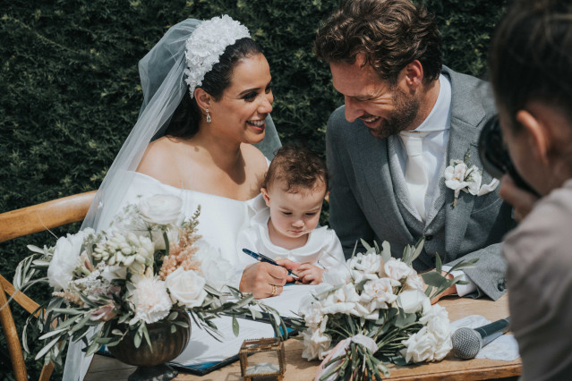 The couple laughs at a table with a small child between them while signing the marriage certificate.