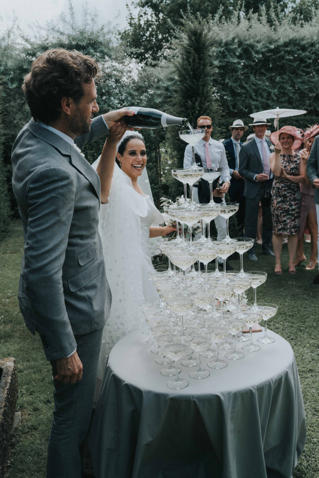 The groom pours champagne over a tower of glasses as the bride and guests watch.