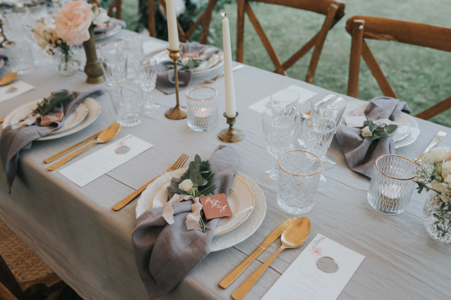 A set table with grey linen napkins, gold cutlery, candles, and small floral accents.