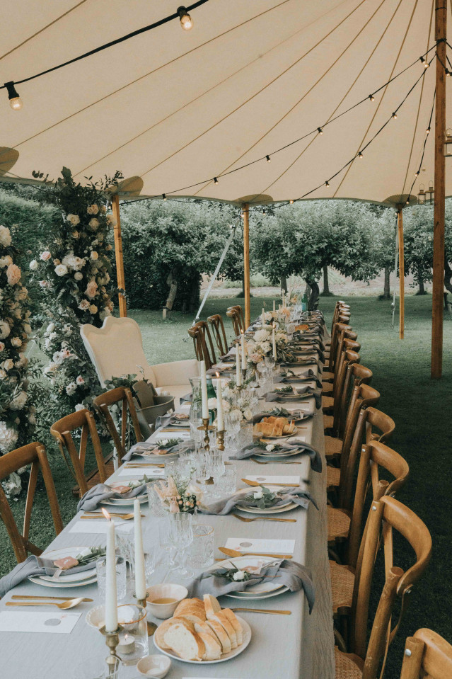 A long set dinner table under a party tent with string lights, flowers, and wooden chairs.