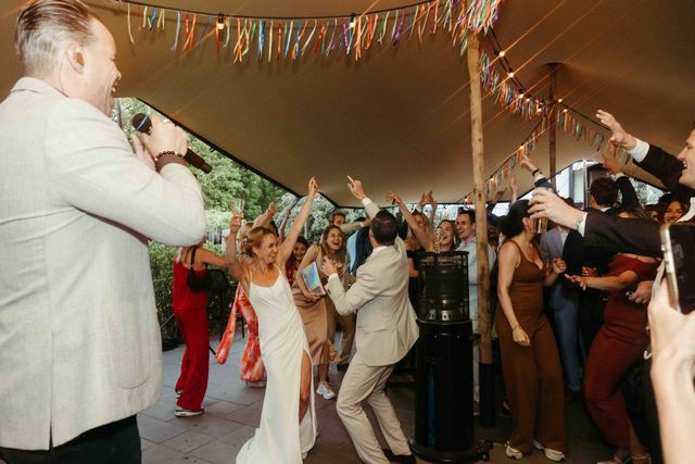 Bride and groom dance with guests under a festive tent decorated with colorful bunting.