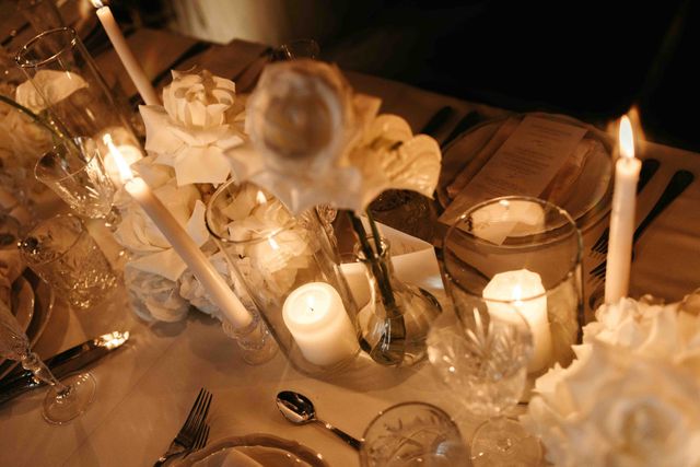 An elegantly set wedding table with white roses, tall candles, crystal glasses, and golden cutlery on a white linen tablecloth.