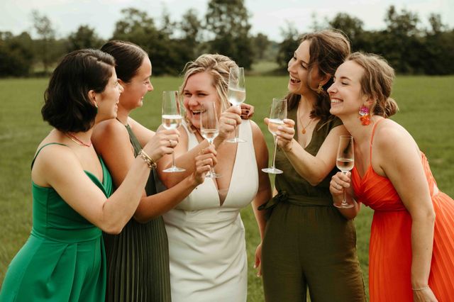 A bride and her friends clink champagne glasses and laugh together outdoors in colorful dresses.