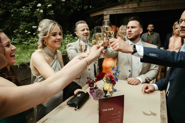 A group of friends toast with champagne glasses, laughing together beside a flower-topped bar.