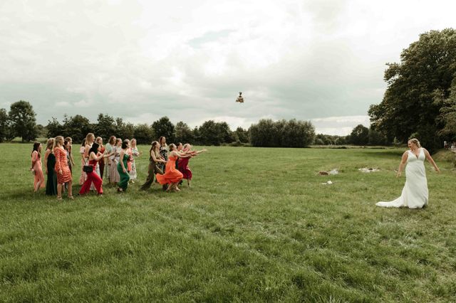 A bride watches her bridesmaids in bright, colorful dresses run forward during the bouquet toss in a green field.