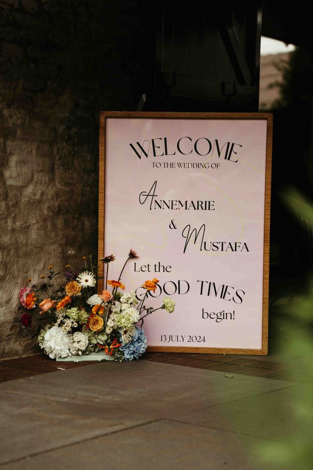 A framed wedding welcome sign decorated with colorful wildflowers at an outdoor venue.