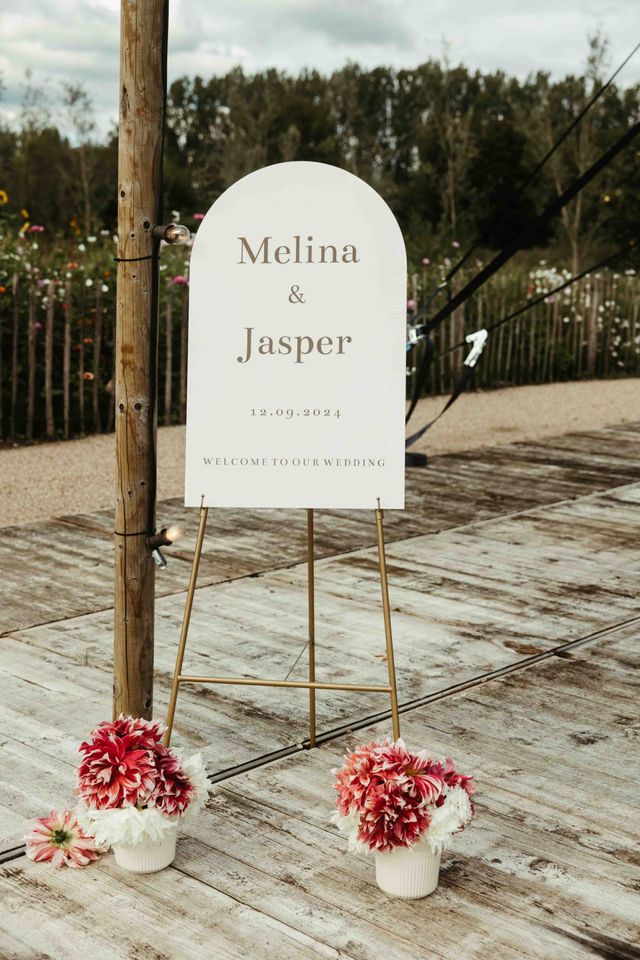 An elegant wedding welcome sign on a gold easel flanked by pink dahlia arrangements at an outdoor venue.