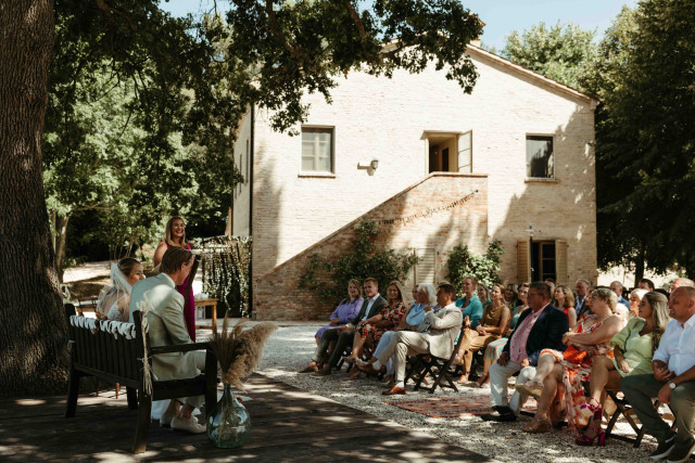 A wide angle view of an outdoor wedding ceremony at a stone farmhouse with guests seated on both sides of the aisle.