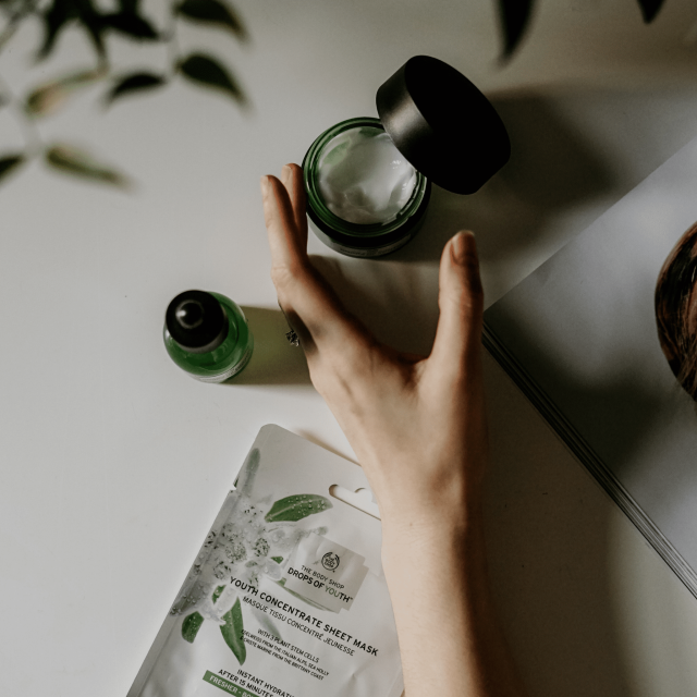 Arm greift nach einem dunkelgruenen Cremetiegel welcher auf einem weissen Untergrund steht