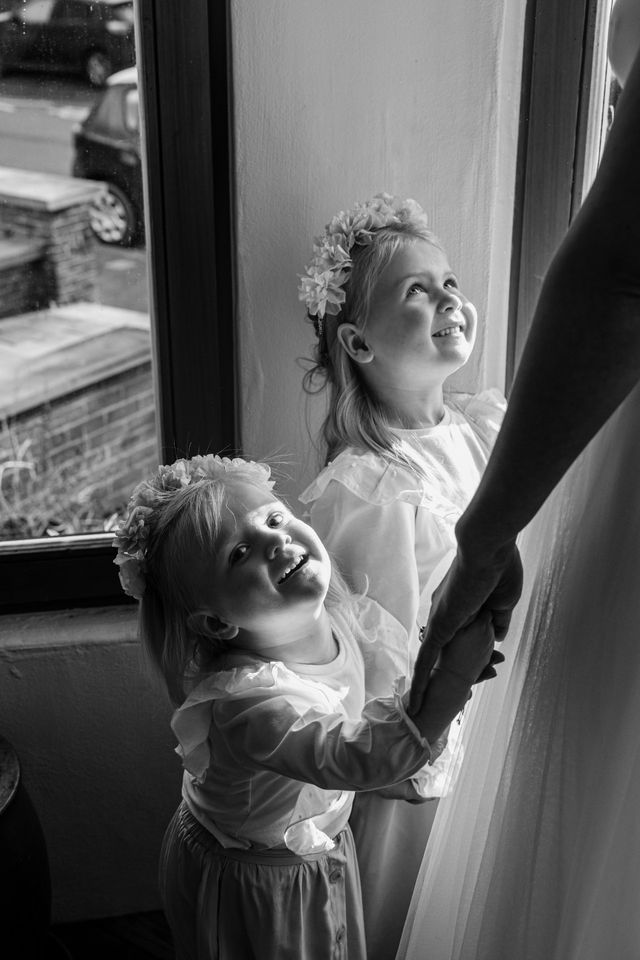 Two laughing flower girls in white dresses with floral crowns hold hands near a window in a black and white photo.