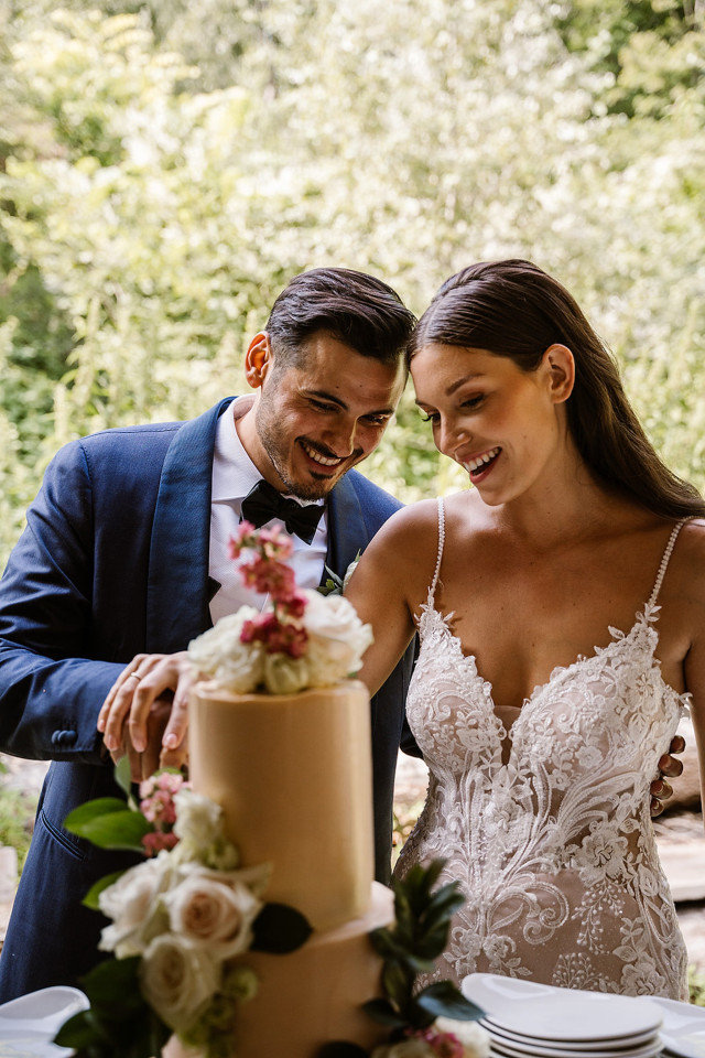 A bride and groom smile together while cutting their tiered wedding cake decorated with roses and greenery.