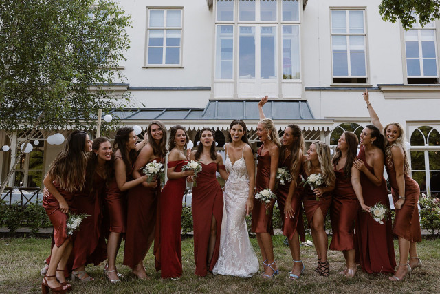 Bridesmaids in matching rust terracotta dresses holding bouquets in front of a white country house.