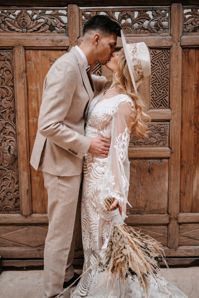 Braut in Boho Brautkleid und Brautstrauß aus Trockenblumen küsst Bräutigam in grauem Anzug