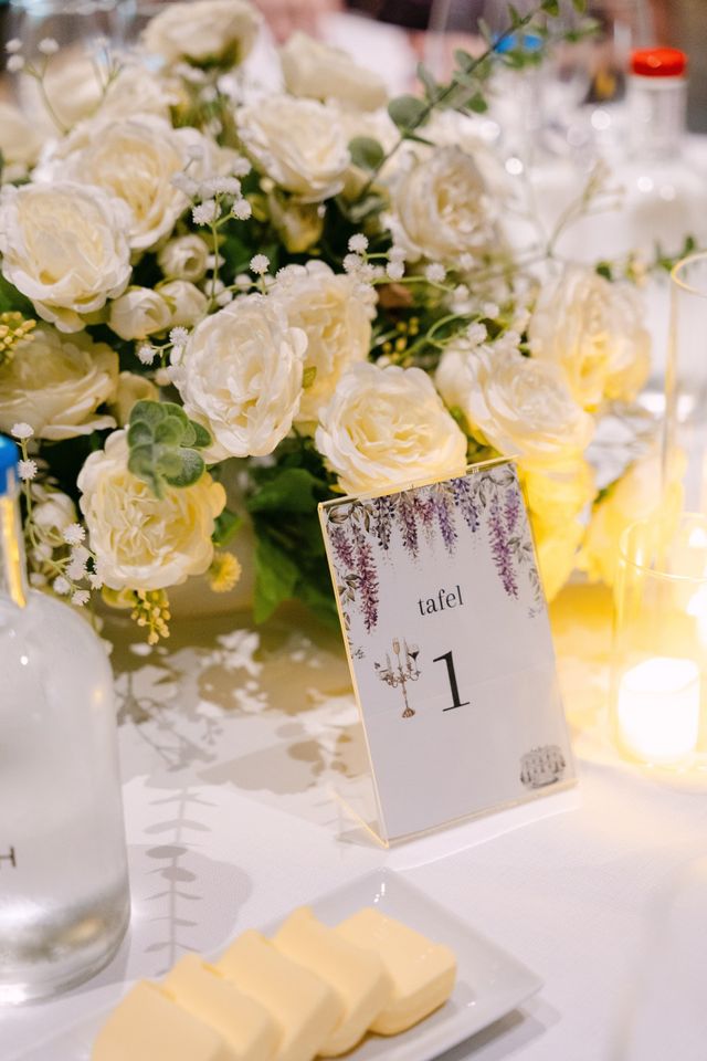 A table number one card with a wisteria illustration beside a lush white rose centerpiece with candles.