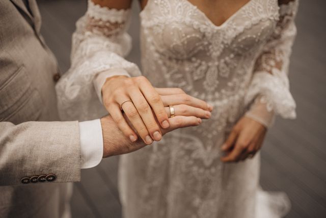 Brautpaar hält Hände übereinander und man sieht die Eheringe und im Hintergrund das Brautkleid
