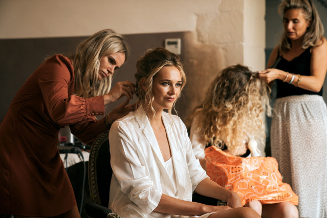 The bride has her makeup and hair done by two stylists during the getting ready morning while a young girl stands nearby.