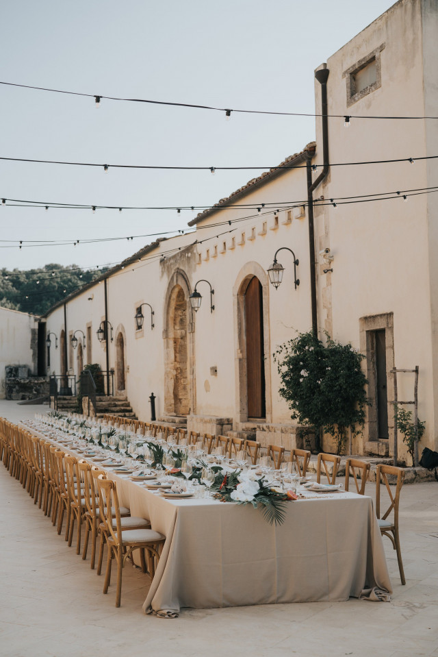 A long banquet table set with white flowers and woven chairs at the reception.