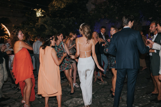 The bride in white trousers with a bare back walks across the party floor.