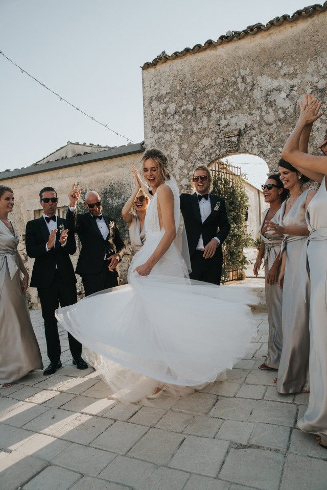 A bride dances and spins her wide dress surrounded by cheering guests on a stone terrace.