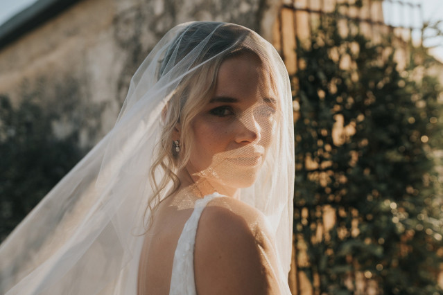 Portrait of the bride with her veil looking over her shoulder in warm sunlight by a stone wall.