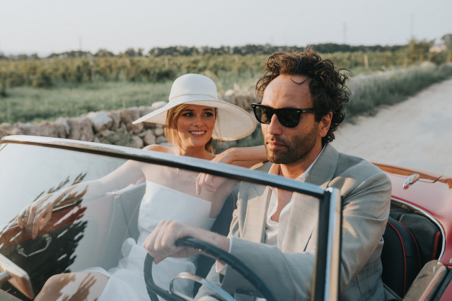A groom in sunglasses drives a red classic convertible while the bride in a hat smiles beside him.