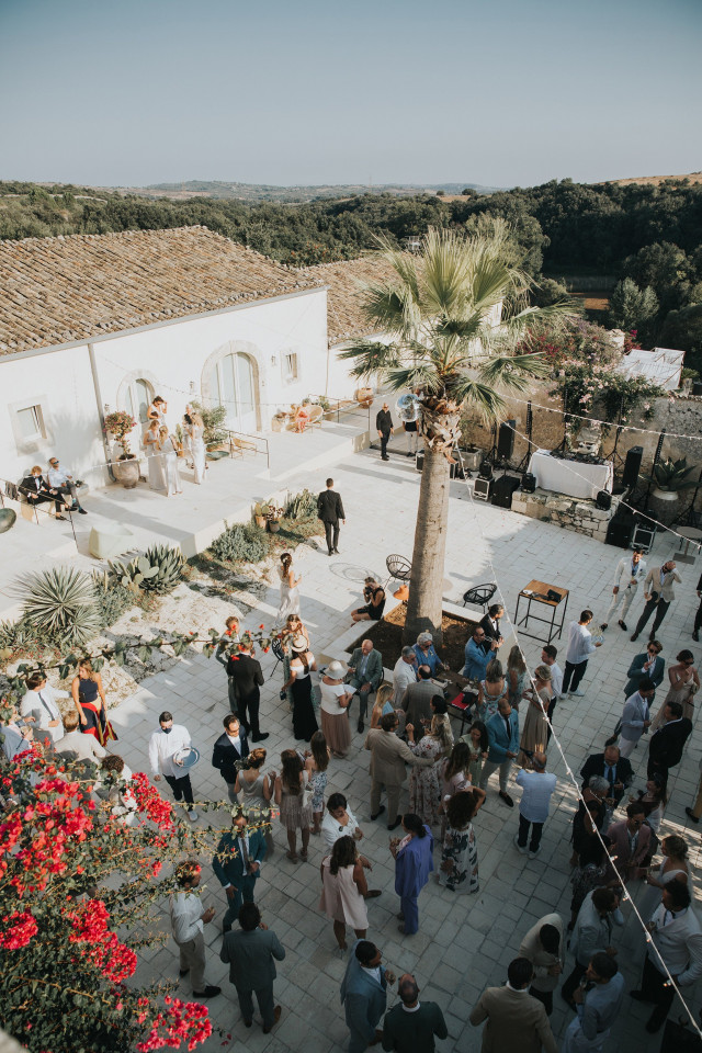 Wedding guests raise their glasses in a toast along a long outdoor dining table adorned with sunflowers in the Italian countryside.