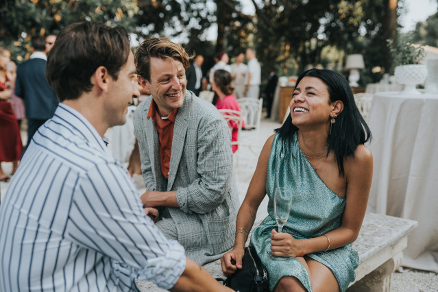 Three guests sit laughing together on a stone bench at the celebration.