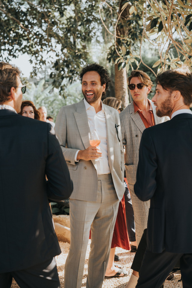 A laughing man in a light grey suit chats with a drink in hand.