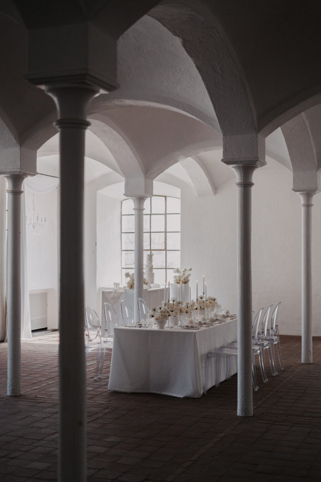 Dekorierter Hochzeitssaal mit Säulen in dem ein langer Hochzeitstisch mit weißen Blumen und weißer Tischdecke aufgebaut ist. Am Tisch stehen durchsichtige Stühle aus Acry.