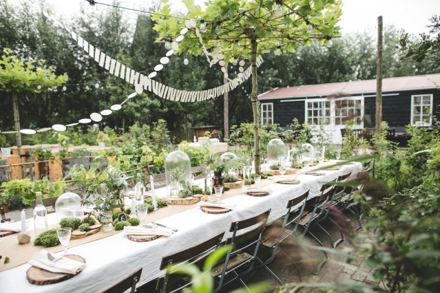 Een elegante buitenbruiloftsreceptie met een lange rustieke tafel vol witte bloemstukken, kaarsen en tafelsettingen onder een natuurlijk bladerdak van groen gebladerte.
