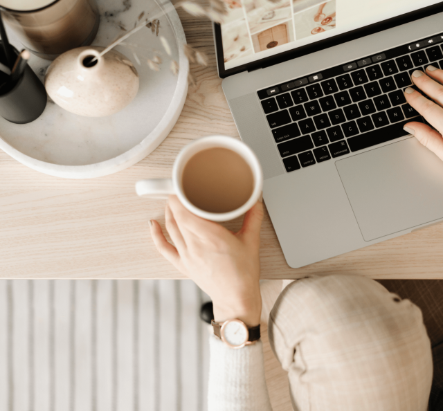 Person am Schreibtisch mit Kaffe in der Hand und einem Laptop
