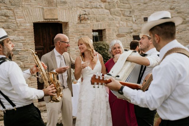 A bride laughs joyfully with musicians and guests outside a stone venue during the reception.