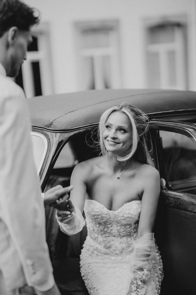 Black and white photo of a bride stepping out of a vintage car as the groom reaches for her hand.