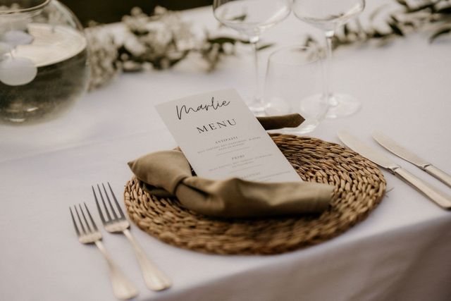 A place setting with a woven placemat, sage linen napkin, handwritten menu card, and glassware, greenery behind.