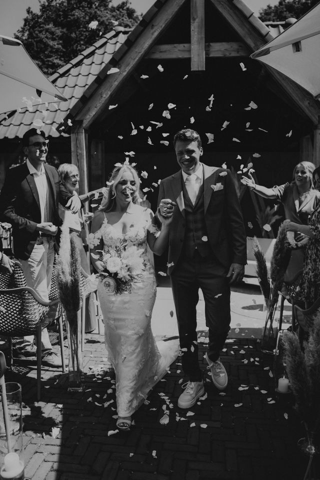 Newlyweds walk joyfully out of the ceremony venue as confetti fills the air, captured in black and white.