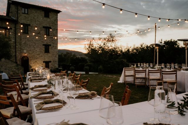 A long al fresco wedding dinner table set on the lawn at sunset, with string lights overhead and a warm pink sky behind a stone villa.