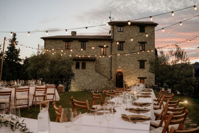 A stone Tuscan villa at sunset with long wedding dinner tables set on the lawn under string lights.