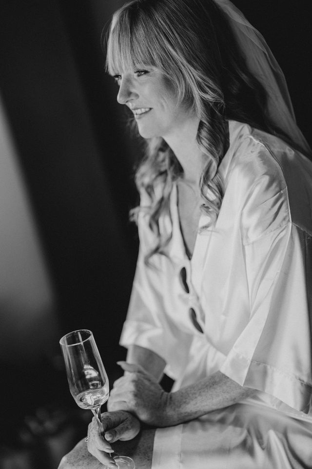 Bride in a white silk robe holding a champagne glass while getting ready
