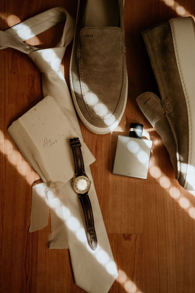 Bovenaanzicht van de accessoires van de bruidegom: suède loafers, een witte stropdas, een horloge en cologne.
