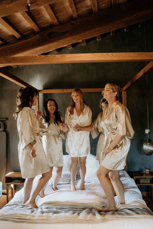 Bridesmaids and the bride in silk robes laugh together on a bed while getting ready.