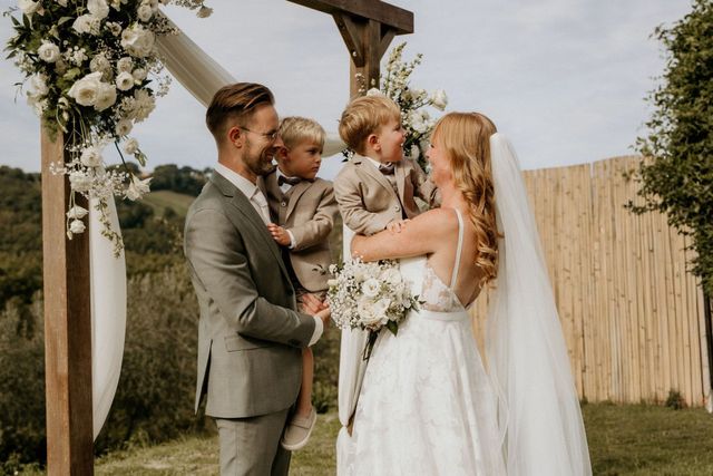 A bride and groom share a tender moment at the flower arch with their two young sons in matching beige suits.