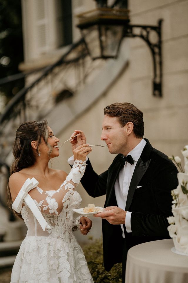 A bride and groom feed each other wedding cake outdoors in front of a chateau, both smiling and laughing.