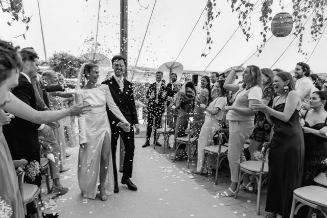 Black and white photo of newlyweds walking back up the aisle through a shower of confetti as guests cheer under a marquee.